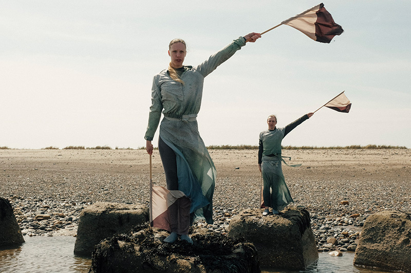 Two performers hold a flag in each of their hands pointing diagonally to their sides. They are stood on large rocks on the beach that are surrounded by water. In the background small stones give way to sand and eventually grass. It is slightly cloudy and sunny.